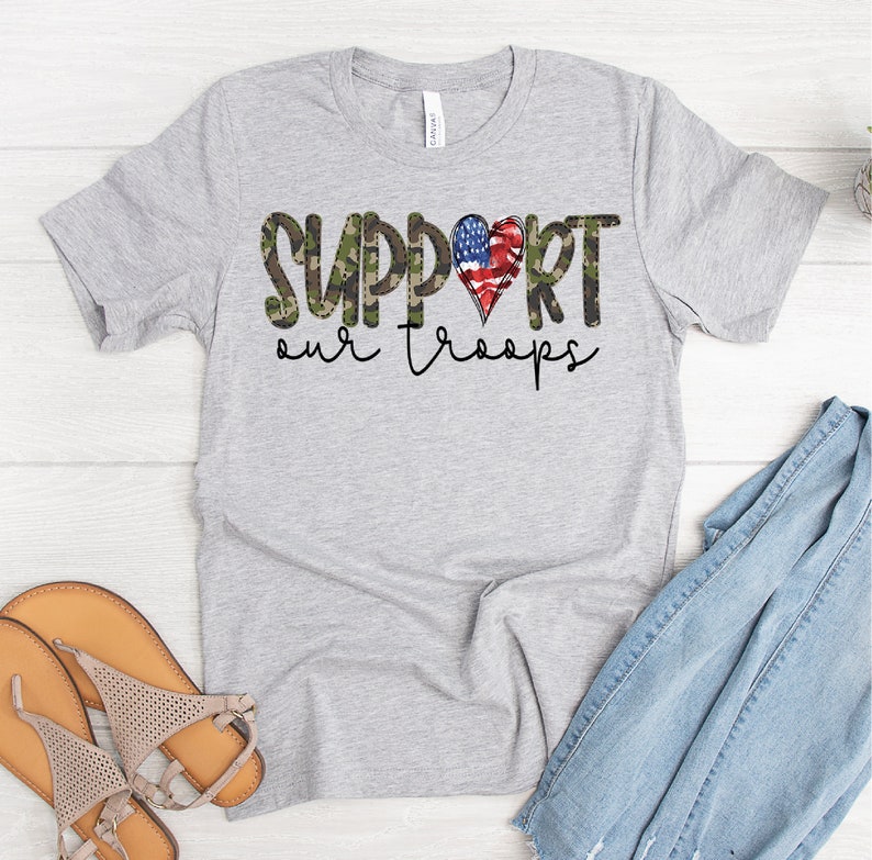 Puede incluir: Una camiseta gris con estampado de camuflaje y un coraz&oacute;n rojo, blanco y azul con las palabras "SUPPORT our troops" impresas en ella.