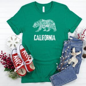 May include: A green t-shirt with a white graphic of a bear made up of California city names. The text "CALIFORNIA" is below the bear. The shirt is laid out on a white wooden surface with a pair of red sneakers, blue jeans, and holiday decorations.