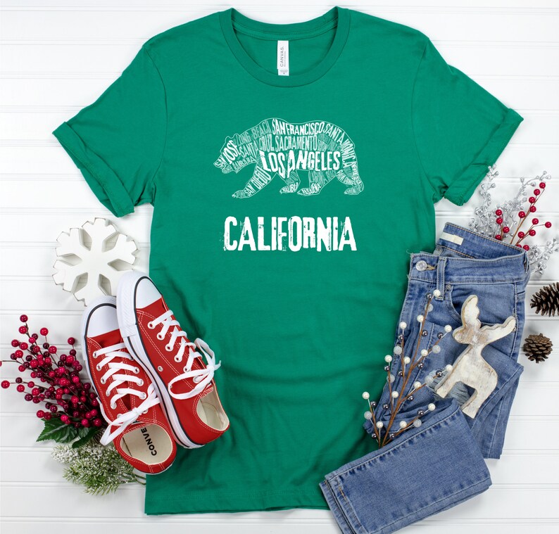 May include: A green t-shirt with a white graphic of a bear made up of California city names. The text "CALIFORNIA" is below the bear. The shirt is laid out on a white wooden surface with a pair of red sneakers, blue jeans, and holiday decorations.