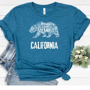 May include: A teal blue t-shirt with a white outline of a California bear. The bear is made up of the names of cities and towns in California, including Los Angeles, San Francisco, and Sacramento. The word "CALIFORNIA" is printed below the bear.