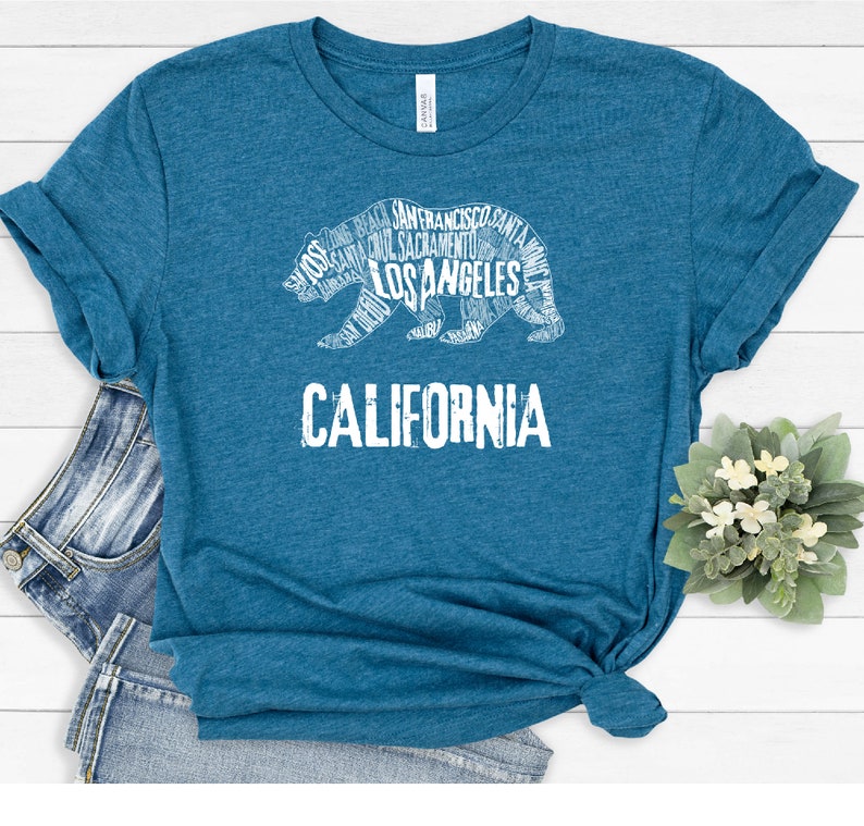 May include: A teal blue t-shirt with a white outline of a California bear. The bear is made up of the names of cities and towns in California, including Los Angeles, San Francisco, and Sacramento. The word "CALIFORNIA" is printed below the bear.