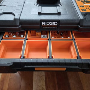 Pode incluir: Caixa de ferramentas preta e laranja RIDGID Pro Gear System Gen 2.0 com v&aacute;rias gavetas retr&aacute;teis. As gavetas s&atilde;o laranja e cont&ecirc;m v&aacute;rias ferramentas e pe&ccedil;as pequenas. A caixa est&aacute; sobre uma superf&iacute;cie de madeira.