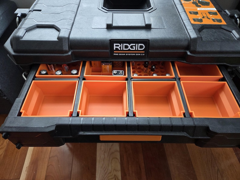 Pode incluir: Caixa de ferramentas preta e laranja RIDGID Pro Gear System Gen 2.0 com v&aacute;rias gavetas retr&aacute;teis. As gavetas s&atilde;o laranja e cont&ecirc;m v&aacute;rias ferramentas e pe&ccedil;as pequenas. A caixa est&aacute; sobre uma superf&iacute;cie de madeira.