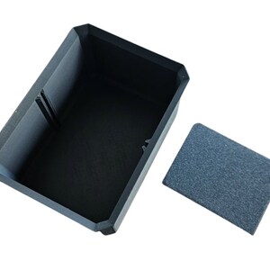 May include: A black rectangular box with a lid. The box is open and the lid is off to the side. The box is made of a textured material.