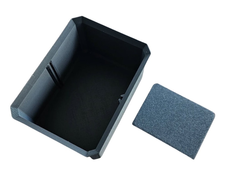 May include: A black rectangular box with a lid. The box is open and the lid is off to the side. The box is made of a textured material.