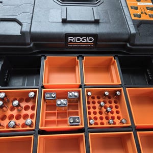 Pode incluir: Caixa de ferramentas preta RIDGID Pro Gear System com compartimentos de armazenamento laranja. As gavetas abertas revelam armazenamento organizado para baterias, incluindo AA, 9V e outros tamanhos. A caixa de ferramentas foi projetada para organiza&ccedil;&atilde;o eficiente.