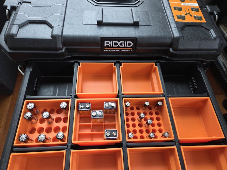 Pode incluir: Caixa de ferramentas preta RIDGID Pro Gear System com compartimentos de armazenamento laranja. As gavetas abertas revelam armazenamento organizado para baterias, incluindo AA, 9V e outros tamanhos. A caixa de ferramentas foi projetada para organiza&ccedil;&atilde;o eficiente.
