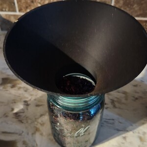 Puede incluir: Un embudo negro se utiliza para verter flores de hibisco secas en un frasco de vidrio transparente con tapa azul.