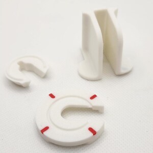 Puede incluir: Tres piezas de pl&aacute;stico blanco, dos tienen forma de C con un peque&ntilde;o espacio en el medio, una es un bloque rectangular con una hendidura en el medio. Las piezas en forma de C tienen marcas rojas.