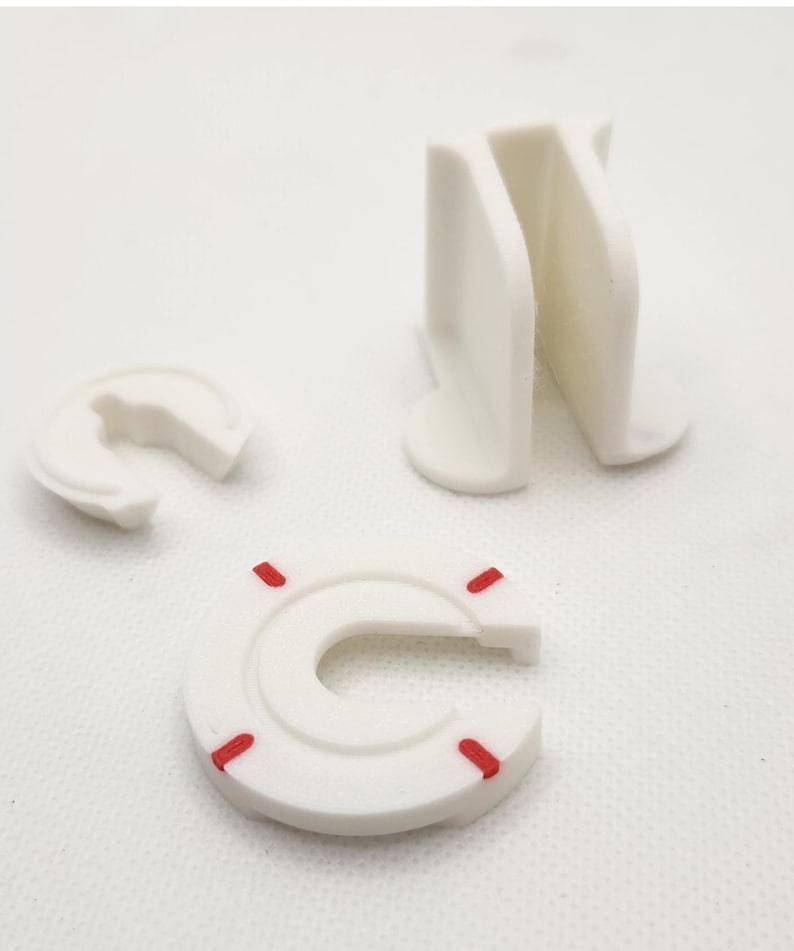 Puede incluir: Tres piezas de pl&aacute;stico blanco, dos tienen forma de C con un peque&ntilde;o espacio en el medio, una es un bloque rectangular con una hendidura en el medio. Las piezas en forma de C tienen marcas rojas.