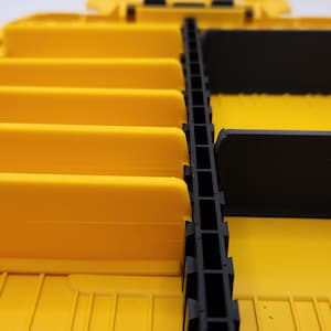 Może przedstawiać: Ż&oacute;łto-czarny plastikowy organizer z wieloma przegr&oacute;dkami. Organizer posiada kilka ż&oacute;łtych przegr&oacute;d i czarne wkładki, przeznaczone do przechowywania drobnych przedmiot&oacute;w. Przezroczysta pokrywa jest częściowo widoczna.