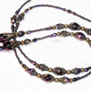 May include: A long necklace featuring dark purple iridescent faceted beads interspersed with small metallic bronze spacer beads and ornate antique bronze bead caps. The design includes a continuous strand of tiny dark purple seed beads.