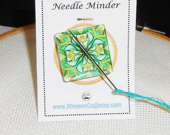 May include: A green and yellow geometric needle minder with a needle threaded with turquoise embroidery floss. The needle minder is on a white card with the text "Needle Minder" and "www.ShivawnCo@etsy.com".