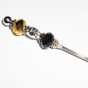 May include: A silver and gold beaded pin with a skull charm and black crystal beads. The pin is attached to a long, thin, silver needle.
