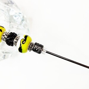 May include: A black and yellow glass bead with a smiley face design, followed by a black bead with silver accents, a white bead with black accents, and a yellow and black glass bead with a swirl design. The beads are attached to a long, thin, silver metal rod.