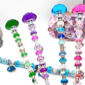 May include: A collection of colorful beaded wands with jewel-toned crystal tops. The wands feature a variety of beads in shades of green, purple, pink, and blue, with silver-colored metal accents. The wands are arranged against a soft, blurred background.