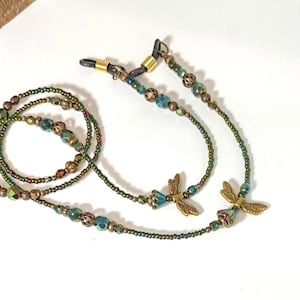 May include: An eyeglass chain featuring small green and iridescent beads, metallic bronze-toned spacers, and two brass-colored dragonfly charms. The chain ends with black rubber loops secured by gold-toned metal coils for attaching to eyewear frames.