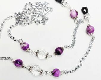 May include: Silver chain eyeglass holder with purple and clear beads. The chain is made of silver-colored metal and features alternating purple and clear glass beads. The eyeglass holder has black rubber loops to attach to glasses.