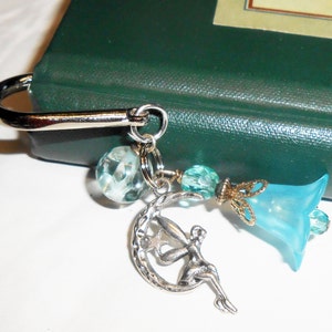May include: A silver bookmark with a blue flower charm, a silver crescent moon charm with a fairy, and a clear glass bead. The bookmark is attached to a green book.