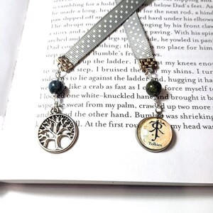 May include: Two handmade bookmarks with gray polka dot ribbon. One bookmark has a tree of life charm and a blue bead. The other has a Tolkien logo charm and a green bead. Both are hanging over an open book.