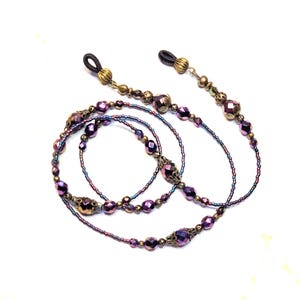 May include: An eyeglass chain featuring iridescent purple and dark blue beads interspersed with antique brass-toned metal spacers and filigree caps. The chain ends in black elastic loops with ribbed brass-toned metal connectors for attachment.