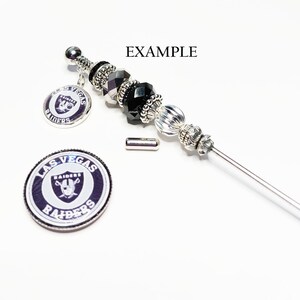 May include: A silver and black beaded pen with a Las Vegas Raiders charm. The pen has black and silver beads, a decorative top, and a charm with the Raiders logo. A separate Raiders logo charm is also shown.