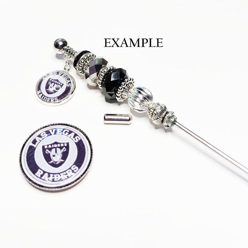 May include: A silver and black beaded pen with a Las Vegas Raiders charm. The pen has black and silver beads, a decorative top, and a charm with the Raiders logo. A separate Raiders logo charm is also shown.