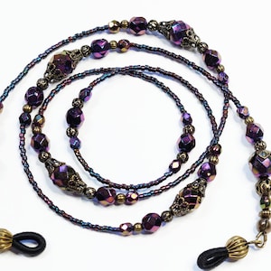 May include: A purple and gold beaded eyeglass chain with decorative metal accents. The chain is made of small beads and larger faceted beads. The chain has two black rubber loops at the ends.