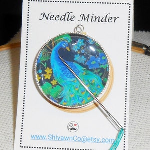 May include: A round needle minder with a peacock design. The needle minder is made of glass and has a silver metal frame. A needle is threaded with teal embroidery floss and is attached to the needle minder.