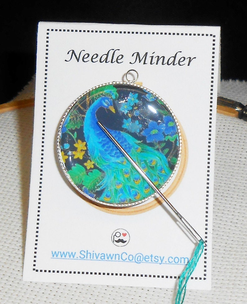 May include: A round needle minder with a peacock design. The needle minder is made of glass and has a silver metal frame. A needle is threaded with teal embroidery floss and is attached to the needle minder.