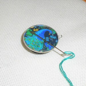 May include: A round, silver-rimmed, glass needle minder with a peacock design. A green thread is threaded through a needle that is sticking out of the minder.
