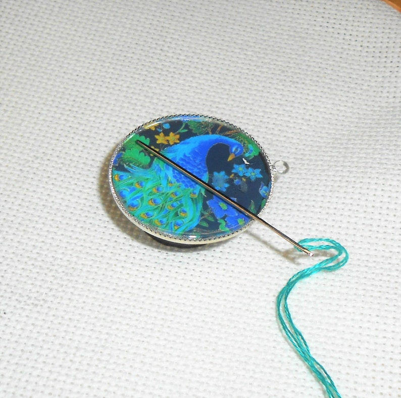 May include: A round, silver-rimmed, glass needle minder with a peacock design. A green thread is threaded through a needle that is sticking out of the minder.