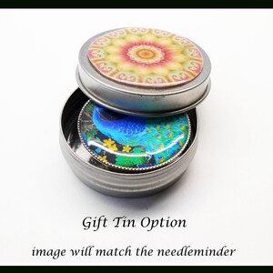 May include: A small silver tin with a round lid. The lid has a colorful mandala design. The tin is open and a round, blue peacock design is visible inside. The text "Gift Tin Option" and "image will match the needle minder" is below the tin.
