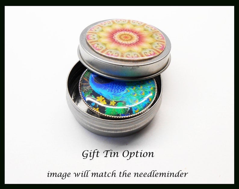 May include: A small silver tin with a round lid. The lid has a colorful mandala design. The tin is open and a round, blue peacock design is visible inside. The text "Gift Tin Option" and "image will match the needle minder" is below the tin.