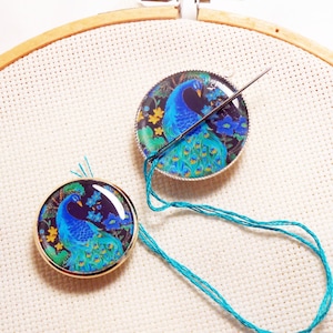 May include: Two round, gold-rimmed, glass ornaments with a peacock design. The ornaments are on a white canvas background. One ornament has a needle threaded through it.