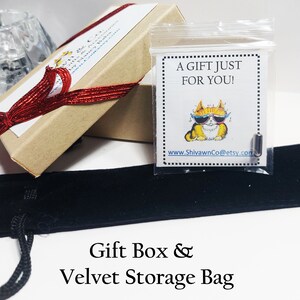 May include: A brown gift box with a red ribbon tied around it. The box is open and a small clear bag with a cat wearing sunglasses and the text "A gift just for you!" is inside. The box is sitting on a black velvet storage bag.