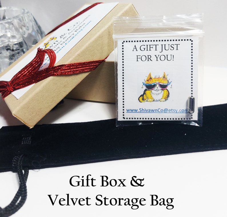 May include: A brown gift box with a red ribbon tied around it. The box is open and a small clear bag with a cat wearing sunglasses and the text "A gift just for you!" is inside. The box is sitting on a black velvet storage bag.