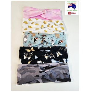 May include: Five fabric headbands with twist-front designs stacked vertically. The patterns include pink camouflage, gold croissants on white, light blue with Australian animals, black with white and yellow butterflies, and a grey camouflage print.