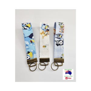 May include: Three fabric key fobs with cartoon character designs. Each key fob has a metal ring and a bronze-coloured metal clasp. One is light blue with cartoon characters, one is white with a pineapple, and one is light blue with cartoon characters.
