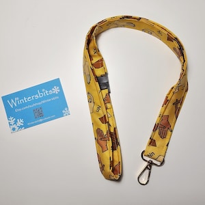 May include: A yellow fabric lanyard featuring a repeating pattern of hands performing American Sign Language letters, including Nn and Xx, with a black plastic breakaway clasp and a gold-toned metal swivel lobster claw clasp at the bottom end.