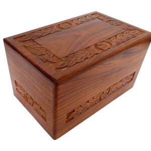 May include: A wooden cremation urn with a carved floral design. The urn is a rich brown colour and has a rectangular shape.