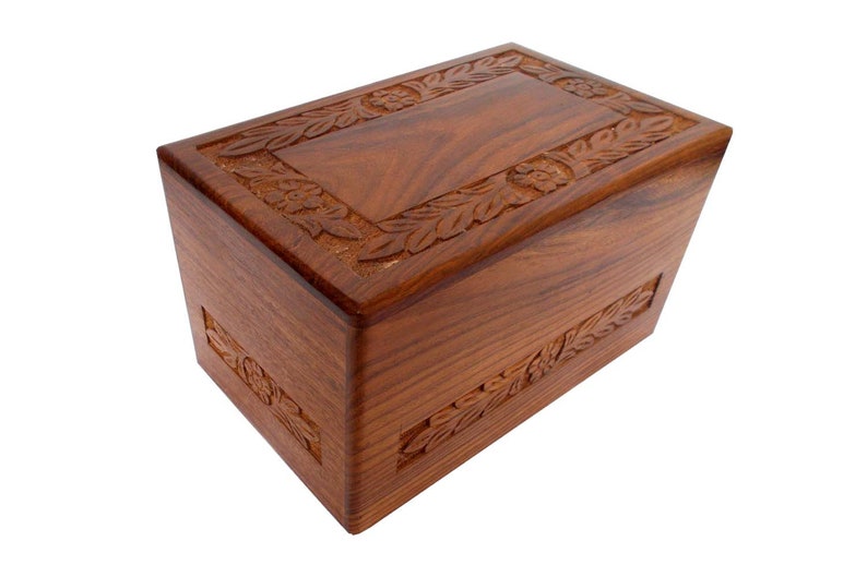 May include: A wooden cremation urn with a carved floral design. The urn is a rich brown colour and has a rectangular shape.