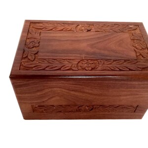 May include: A dark brown wooden cremation urn with intricate floral carvings around the top and bottom edges.