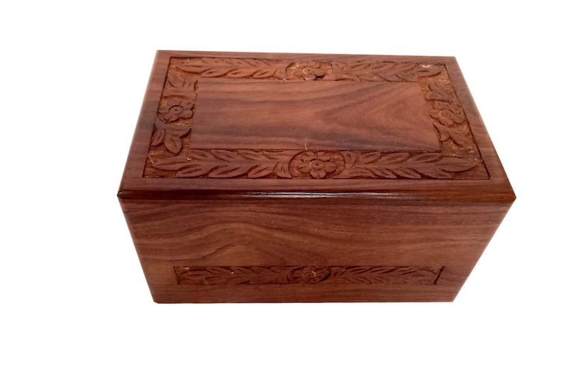 May include: A dark brown wooden cremation urn with intricate floral carvings around the top and bottom edges.