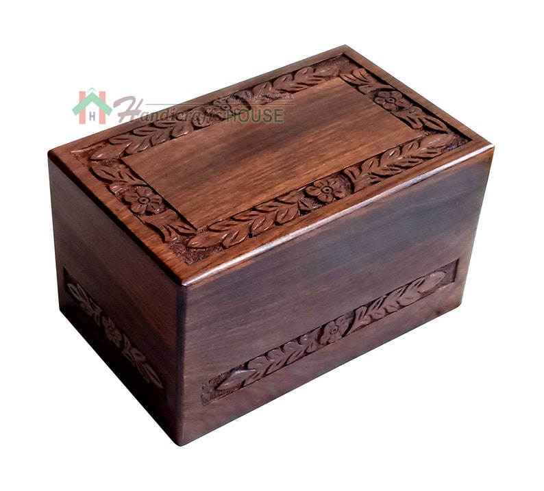 May include: A dark brown wooden box with a carved floral design around the edges. The box has a hinged lid and is made of solid wood.