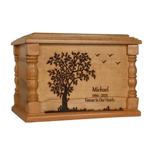 Op de afbeelding: Een lichtbruine houten urn met een geëtste boom en vogels. De tekst luidt "Michael 1956-2025 Forever In Our Hearts". De urn heeft decoratieve pilaren aan de zijkanten en een rechthoekige vorm.
