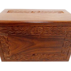 May include: A wooden cremation urn with a carved floral design. The urn is made of dark wood and has a rectangular shape.