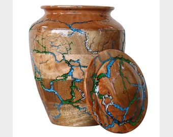 May include: A wooden urn with a lid, featuring a natural wood grain pattern. The urn is adorned with blue and green resin accents, creating a unique, decorative design. The urn is a light brown color.