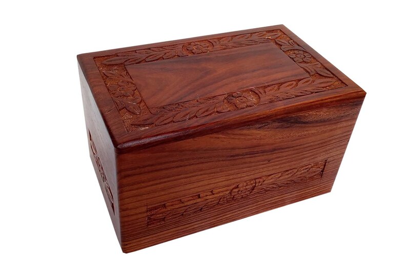 May include: A wooden cremation urn with a carved floral design. The urn is made of dark reddish-brown wood and has a rectangular shape.