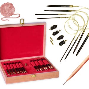 May include: A wooden box with a red velvet interior containing a set of 12 knitting needles. The needles are made of wood and have gold tips. The box is open and the needles are arranged in rows. There is a ball of yarn and a crochet hook in the background.